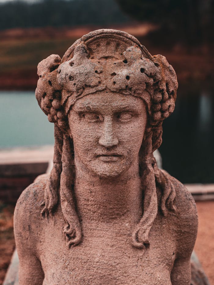 Close-up of a weathered ancient stone sculpture of a woman with intricate headpiece near a serene water body.